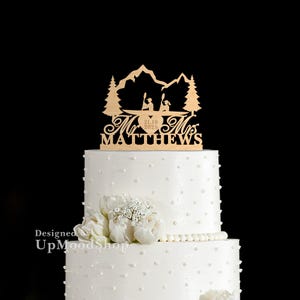 以下が含まれることがあります： 山と木を背景に、カヌーに乗ったカップルが描かれた木製トッパーを飾った白いウェディングケーキ。トッパーには「Mr. & Mrs. Matthews」とハートの中に日付「21.10.2025」が書かれている。