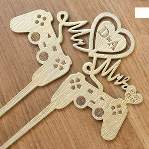 May include: Gold-coloured wooden cake topper featuring two video game controllers, a heart with the initials "D & A" and the word "Mr & Mrs" with the date "02.07.2018".
