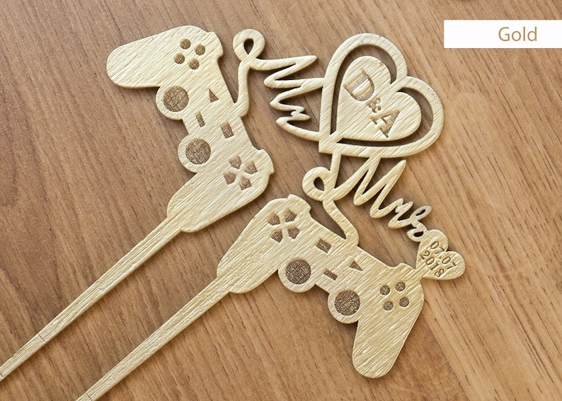 May include: Gold-coloured wooden cake topper featuring two video game controllers, a heart with the initials "D & A" and the word "Mr & Mrs" with the date "02.07.2018".