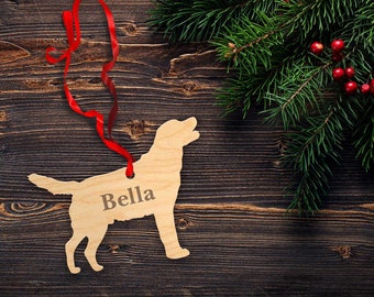 Op de afbeelding: Een houten kerstversiering in de vorm van een hond met de naam "Bella" erop gegraveerd. De versiering hangt aan een rood lint en is omringd door een groene dennentak met rode bessen.