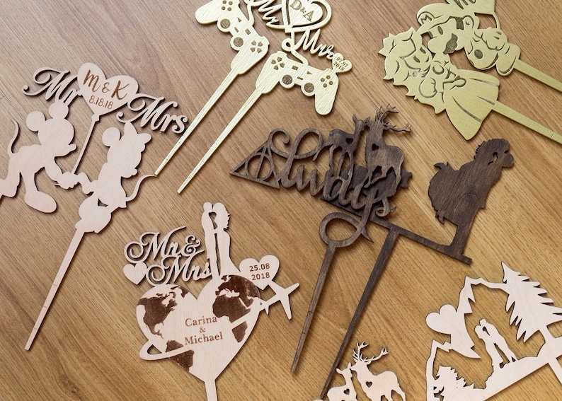 Puede incluir: Surtido de adornos para tartas de madera con diferentes temas, incluyendo una pareja cogida de la mano, una pareja con un globo terr&aacute;queo, una pareja con un mando de videojuegos, una pareja con un coraz&oacute;n, una pareja con un ciervo, una pareja con una monta&ntilde;a y una pareja con un castillo. Los adornos est&aacute;n hechos de madera y est&aacute;n pintados en varios colores.