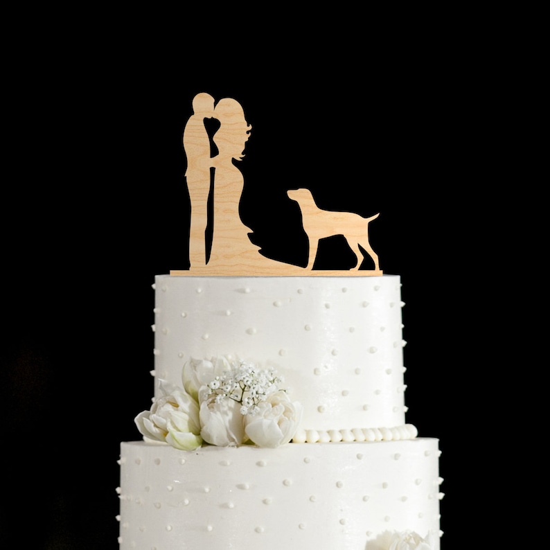 May include: A white wedding cake with a wooden topper of a couple kissing and a dog standing behind them. The cake is decorated with white icing and white polka dots.