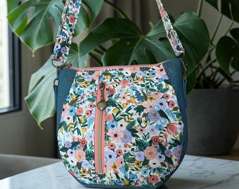 May include: A floral print shoulder bag with a blue cork base and a pink zipper. The bag features a floral pattern with pink, blue, and yellow flowers. The strap is also floral, and the zipper pull is heart-shaped.