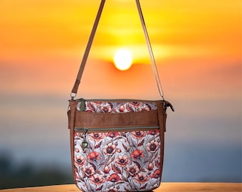 May include: A crossbody bag featuring a floral pattern of red and orange poppies on a light background, accented with brown cork-like trim. The bag includes two zippered compartments with green zippers and a small heart-shaped charm on the front zipper.