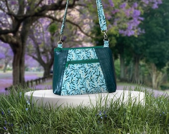 May include: A green and blue floral patterned crossbody bag with a green faux leather trim and adjustable strap. The bag is sitting on a white platform in front of a green grassy area.