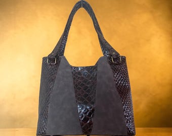 May include: A brown tote bag with a crocodile-textured exterior and suede-like panels. The bag has a rectangular shape with a structured design and long handles. The bag is sitting on a wooden surface.