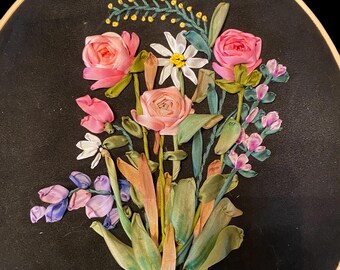 May include: A close-up of a floral embroidery hoop art piece. The hoop is filled with pink and white silk ribbon flowers, green leaves, and purple and blue ribbon accents. The background is black.