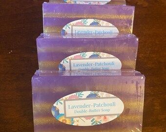 May include: Four bars of Lavender-Patchouli Double-Butter Soap are stacked. The soap has a lavender and gold color scheme. Each bar is wrapped in clear plastic and has an oval label with the product name and floral accents.