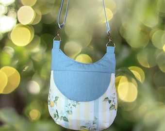 May include: A light blue crossbody bag with a curved top flap. The bag features a floral and striped pattern in yellow, white, and light blue. The bag has a light blue strap and metal hardware.