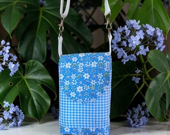 May include: A small, blue floral and gingham print crossbody bag with a white strap. The bag features a flap closure and a white strap with silver-tone metal clasps. The background includes green leaves and blue flowers.