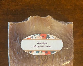 May include: A bar of amethyst cold process soap, wrapped in clear plastic. The soap is a light brown color with a textured surface. A white oval label with floral accents and the text "Amethyst cold process soap" is affixed to the front.