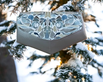 May include: A gold-colored purse with a chain strap and a blue and brown patterned flap. The purse is hanging from a snowy evergreen tree branch.