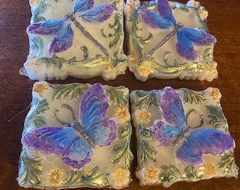 May include: Four decorative square soaps with butterfly and dragonfly designs. Each soap features a light green background with gold accents and a detailed insect in shades of blue and purple. The soaps are arranged on a wooden surface.