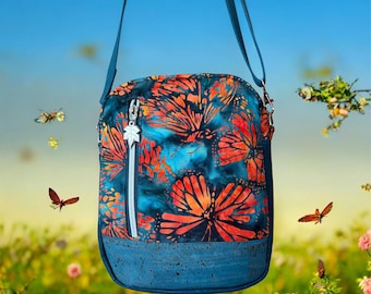 May include: A blue crossbody bag with a vibrant butterfly pattern in orange and blue. The bag has a blue cork base, a silver zipper, and a leaf-shaped zipper pull. The bag is suspended by a blue strap.