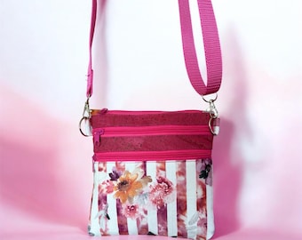 May include: A crossbody bag featuring a magenta strap and a top section made of textured dark pink material. The lower portion displays a pattern of vertical white stripes overlaid with a floral design in shades of orange, pink, and brown.