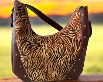 May include: A brown leather hobo bag with a zebra print design and gold-toned studs. The bag has a long strap and a zipper closure.