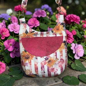 May include: A floral crossbody bag with a red cork pocket. The bag features a white and red striped pattern with watercolor-style flowers in shades of orange, yellow, and purple. The strap and top of the bag have a matching floral design.