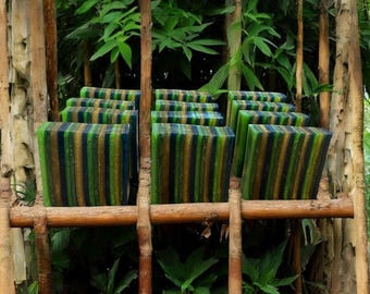 May include: A collection of handmade, multi-colored, striped bars of soap arranged on a wooden rack. The soap is green, blue, and gold with a shimmery finish. The soap is displayed in a natural setting with green foliage.