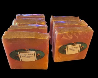 May include: Several bars of mango-peach cold process soap, wrapped in clear plastic. The soap has a gradient of orange and pink hues. Each bar has a label with the product name and a tropical leaf design.