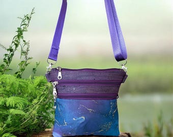 May include: A crossbody bag featuring a purple cork upper section and a blue lower section with a gold marble pattern. The bag includes two horizontal zippered compartments, silver-toned metal zipper pulls, and an adjustable purple fabric strap.