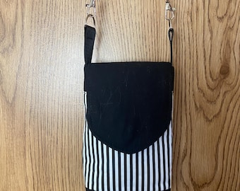 May include: Black and white striped crossbody bag with a black flap and strap. The bag has a rectangular shape with a curved flap closure and a long, adjustable strap. The bag is hanging against a wooden background.