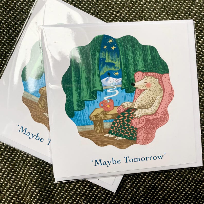 May include: A white bear sits in a pink armchair with a green blanket and a red mug of hot drink. The bear is looking out a window at a snowy scene with a green curtain and a starry night sky. The text 'Maybe Tomorrow' is written below the image.