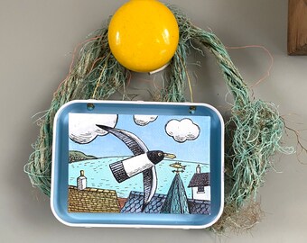 Art in a tin hanging decoration - Gull over the rooftops of a Cornish style harbour.