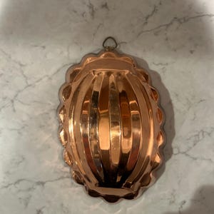 May include: A copper-colored, antique-style mold with a scalloped edge and a loop for hanging. The mold has a ribbed design and a shiny finish, reflecting light. The overall shape is oval, and it appears to be made of metal.