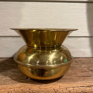 May include: A small, round, brass vase with a flared top. The vase has a smooth, polished finish.