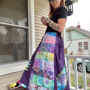 May include: A long, purple wrap skirt with a patchwork design featuring floral patterns in various colors. The skirt has a tie closure and a decorative floral design on the side. The skirt is worn outdoors.