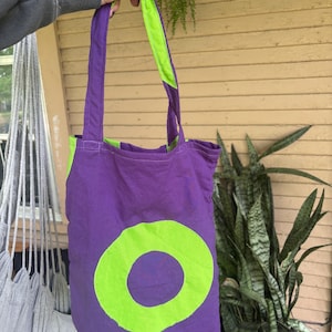 May include: A purple tote bag with a large, lime green circle design. The bag has lime green accents on the straps and interior. The bag is made of fabric and is being held up by a person.