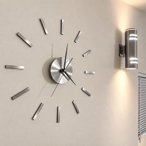 May include: A wall-mounted clock featuring a circular brushed silver center hub with three thin black hands. Twelve individual rectangular silver markers are arranged in a circle around the central hub to indicate the hours.