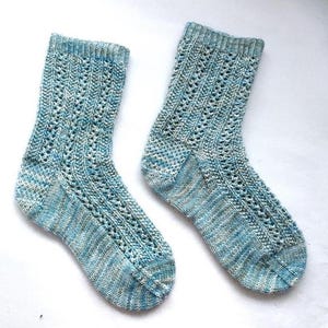 Può includere: Un paio di calzini lavorati a mano nei toni del blu, bianco e grigio. I calzini presentano un delicato motivo traforato e polsini a coste. Il filato ha un sottile effetto variegato, che aggiunge profondità al design. Questi calzini sono perfetti per l'uso quotidiano.