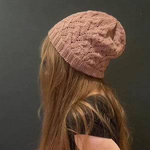 Può includere: Un berretto lavorato a maglia di colore rosa antico. Il cappello presenta un design traforato strutturato. Il berretto è realizzato con un filato morbido, forse lana. Il cappello è mostrato di lato.