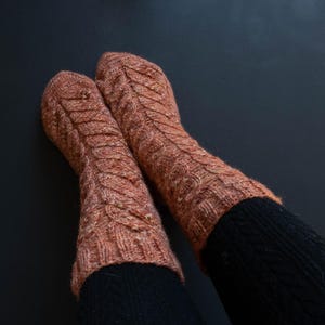 May include: A pair of knitted socks in a warm, reddish-brown hue with a cable knit design. The socks are worn over black leggings, creating a contrast. The socks appear soft and cozy, perfect for cold weather.
