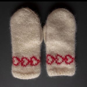 Può includere: Un paio di guanti in maglia bianco sporco con un motivo a cuore rosso sui polsini. I guanti sono realizzati in un filato morbido e strutturato, progettati per tenere le mani al caldo. Lo sfondo è grigio scuro.