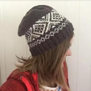 Può includere: Un berretto lavorato a maglia grigio scuro con un motivo geometrico bianco. Il cappello è morbido e sembra fatto a mano. Il berretto è indossato da una persona che indossa una giacca rossa.