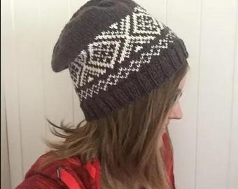 May include: A dark gray knit beanie with a white geometric pattern. The hat is slouchy and appears to be handmade. The beanie is worn by a person wearing a red jacket.