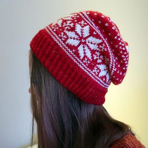 Può includere: Un berretto di lana rosso e bianco con un motivo a fiocco di neve. Il berretto è indossato sulla testa di una persona.
