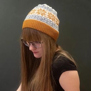 Può includere: Un berretto lavorato a maglia con un bordo giallo senape, una sezione centrale grigia e bianca fantasia e una parte superiore bianca. Il cappello presenta un motivo a fiocco di neve. Il cappello è realizzato in un materiale morbido e caldo, ideale per il freddo.