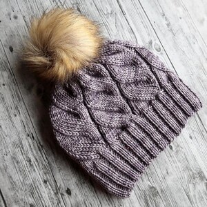 Può includere: Un cappello lavorato a maglia viola con un motivo a trecce e un grande e soffice pompon color marrone chiaro. Il cappello è realizzato in filato strutturato, progettato per calore e stile. Un accessorio invernale.