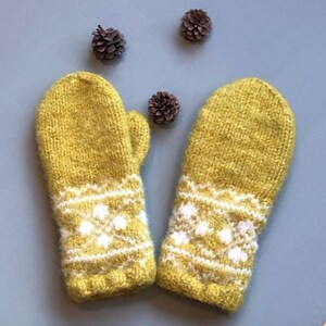 Könnte beinhalten: Senfgelbe Strickfäustlinge mit weißem Schneeflocken- und Streifenmuster. Die Fäustlinge werden auf einer blauen Oberfläche mit drei kleinen Tannenzapfen gezeigt.