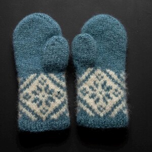 May include: A pair of blue knitted mittens with a white geometric pattern on the lower portion. The mittens are made of a textured yarn and are designed for warmth and comfort. The background is dark.