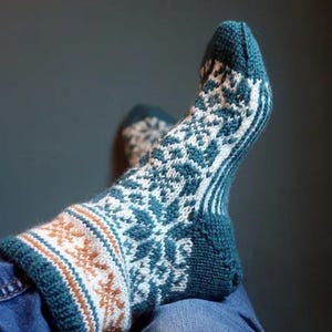 May include: A knitted sock with a teal, white, and brown geometric pattern. The sock is on a person's foot, with the cuff featuring a brown and white design. The sock's main body has a teal and white snowflake-like pattern.