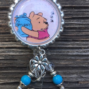 May include: A Winnie the Pooh themed badge reel with a silver bottle cap featuring Winnie the Pooh holding a honey pot. The badge reel has a silver flower charm and two turquoise beads.