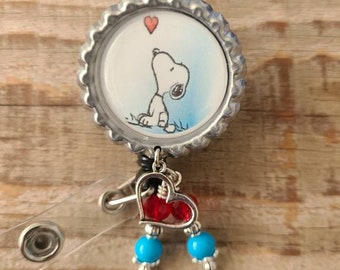 May include: A Snoopy-themed retractable badge reel. The image features Snoopy looking up at a heart. The reel has a silver bottle cap design, a heart charm with red beads, and blue beads. The background is a wooden surface.