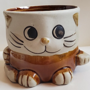 May include: A ceramic mug shaped like a cat with brown and white glaze. The cat has a brown nose, black eyes, and a white belly.
