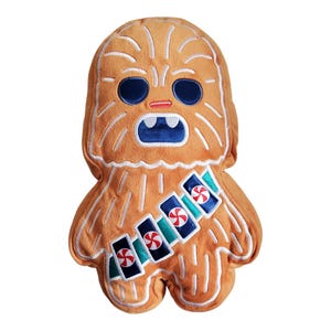 Op de afbeelding: Een pluche Chewbacca kussen met een gingerbread man ontwerp. Het kussen is bruin en wit met een blauw en groen pepermuntpatroon.