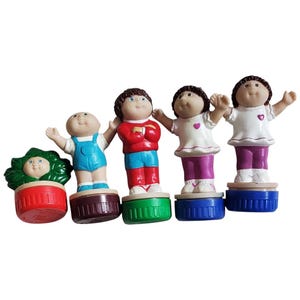 Könnte beinhalten: Fünf Cabbage Patch Kids Figuren, jede mit einem anderen Outfit und einer anderen Pose, stehen auf bunten Sockeln. Die Figuren sind aus Kunststoff und haben bemalte Details. Die Sockel sind aus Kunststoff und haben eine glatte Oberfläche.