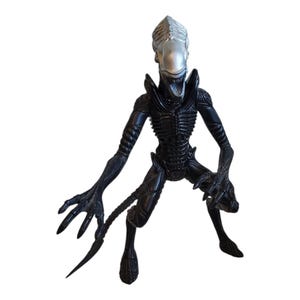 Puede incluir: Figura de acción Xenomorfo negra con cabeza plateada. La figura tiene una estructura esquelética detallada, brazos largos con manos con garras y una cola segmentada. La figura está en posición agachada.